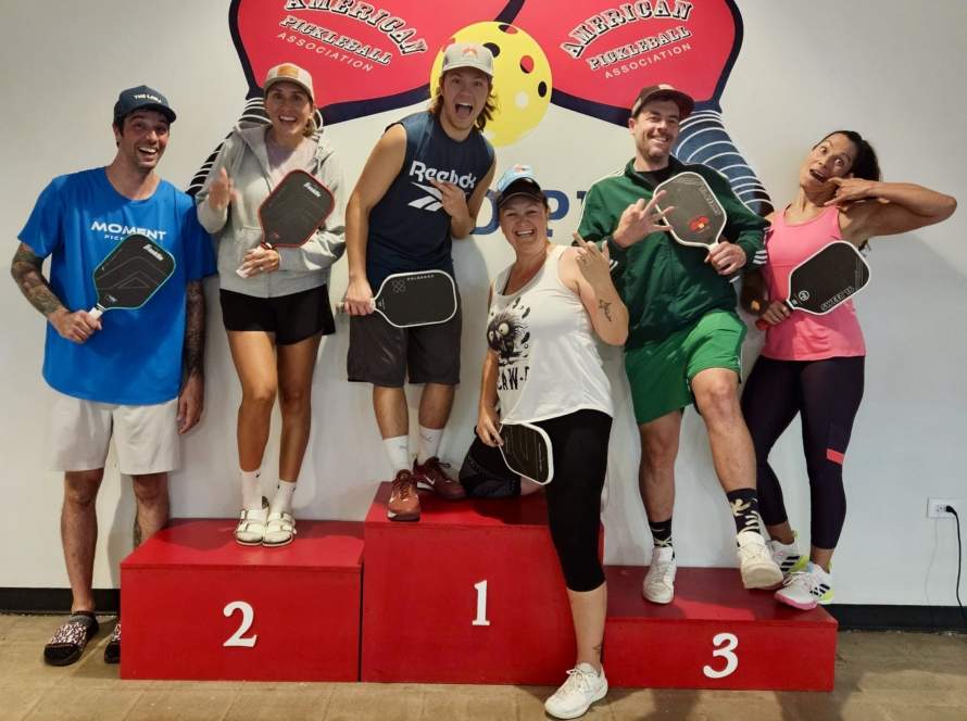 A lineup of pairs of male and female players on podiums numbered one, two and three. These players are very animated. Pickleball Food Pub